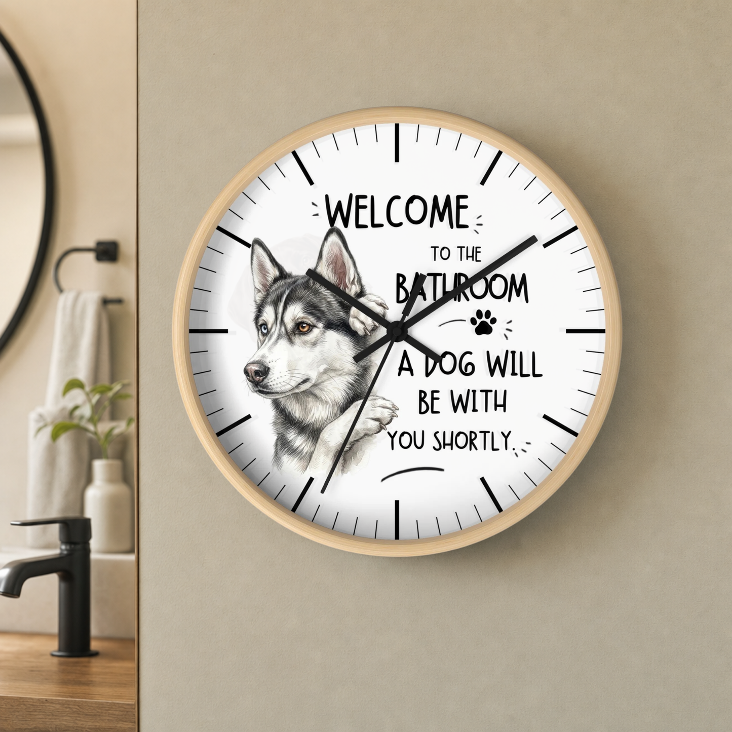 A round wall clock with a light wood frame hangs on a beige wall in a bathroom setting. The clock face is white with black text reading 'WELCOME TO THE BATHROOM A DOG WILL BE WITH YOU SHORTLY' and a detailed husky illustration on the left side.