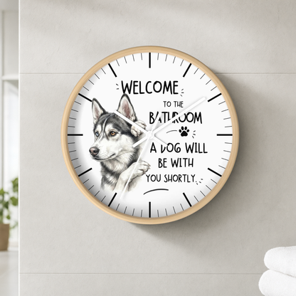 A round wall clock with a light wood frame and a white face, featuring a husky illustration and the text 'WELCOME TO THE BATHROOM A DOG WILL BE WITH YOU SHORTLY'. The clock is mounted on a light gray tiled wall.