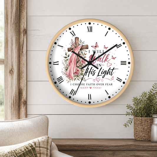 A round wall clock with a light wood frame hangs on a white shiplap wall above a wooden table. The clock face is white with black Roman numerals, black hands, and a central religious illustration featuring a butterflies in pink and a wooden cross.