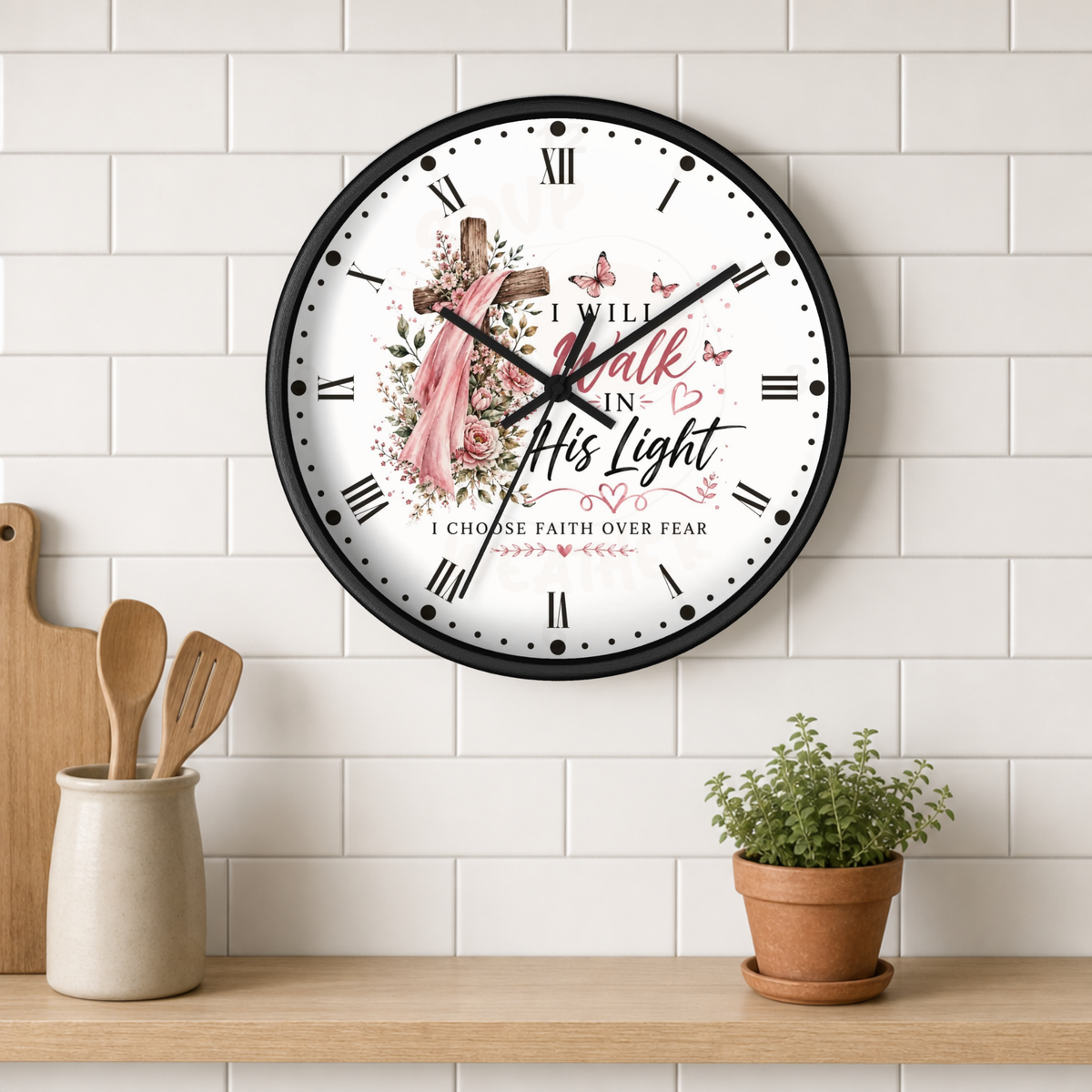 A round wall clock with a black frame and a white face featuring a floral cross, butterflies, and the text 'I WILL walk in His light I CHOOSE FAITH OVER FEAR'. The clock is shown hanging on a white subway tile wall above a wooden shelf with kitchen utensils and a potted plant.