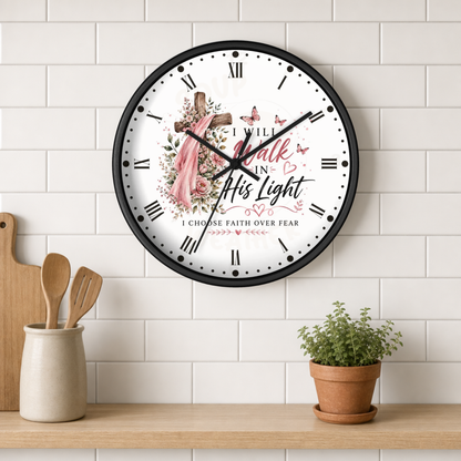 A round wall clock with a black frame and a white face featuring a floral cross, butterflies, and the text 'I WILL walk in His light I CHOOSE FAITH OVER FEAR'. The clock is shown hanging on a white subway tile wall above a wooden shelf with kitchen utensils and a potted plant.