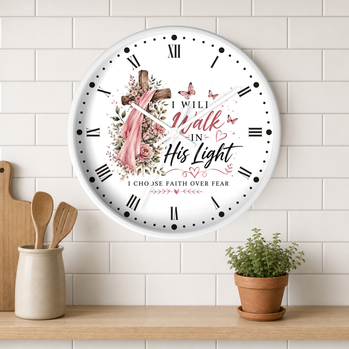 A round white wall clock with Roman numerals and a Christian-themed floral design featuring a cross, pink veil, butterflies, and the text 'I Will Walk In His Light' and 'I Choose Faith Over Fear'. The clock is mounted on a white subway tile wall above a wooden shelf with a small potted plant and wooden utensils.