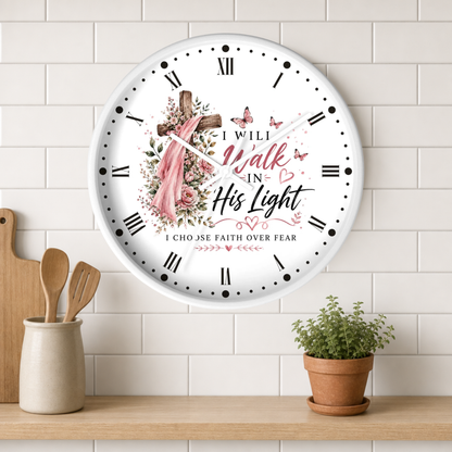 A round white wall clock with Roman numerals and a Christian-themed floral design featuring a cross, pink veil, butterflies, and the text 'I Will Walk In His Light' and 'I Choose Faith Over Fear'. The clock is mounted on a white subway tile wall above a wooden shelf with a small potted plant and wooden utensils.