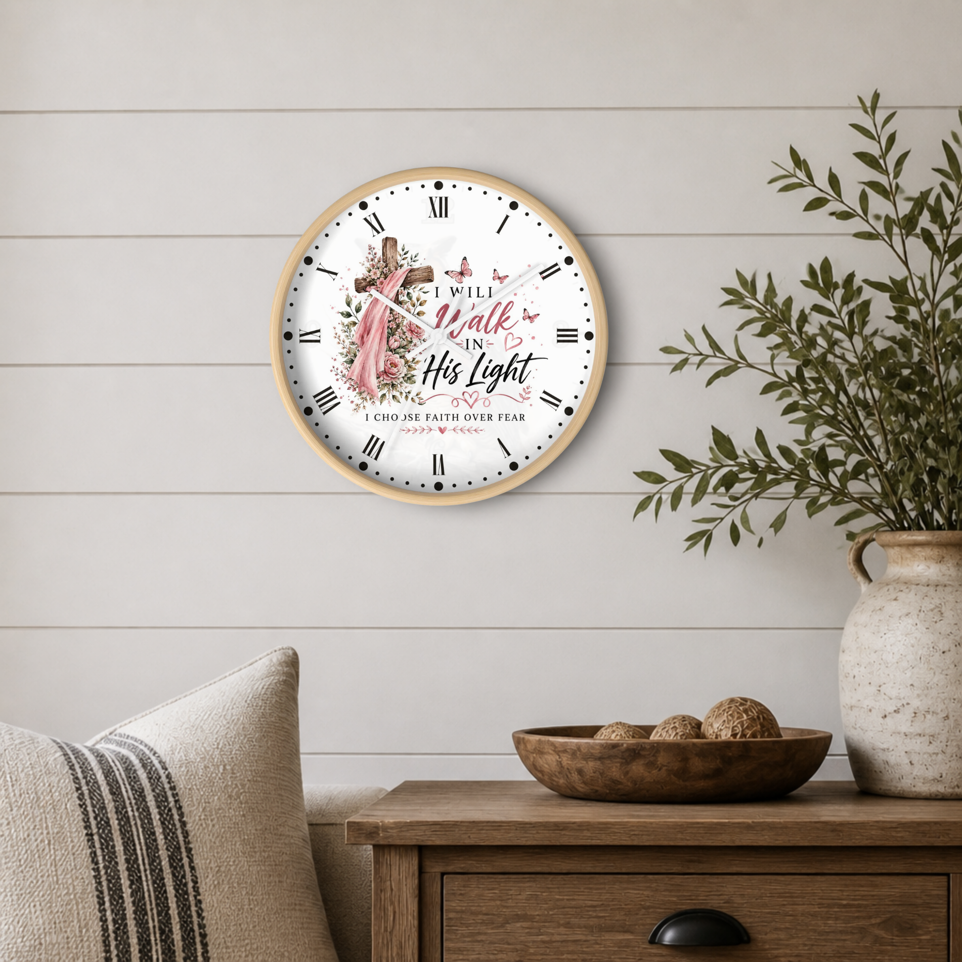 A round wall clock with a light wood frame hangs on a white shiplap wall above a wooden side table. The clock face is white with black Roman numerals, a central illustration of a pink butterflies and a cross, and the text 'I Will Walk In His Light' and 'I Choose Faith Over Fear.