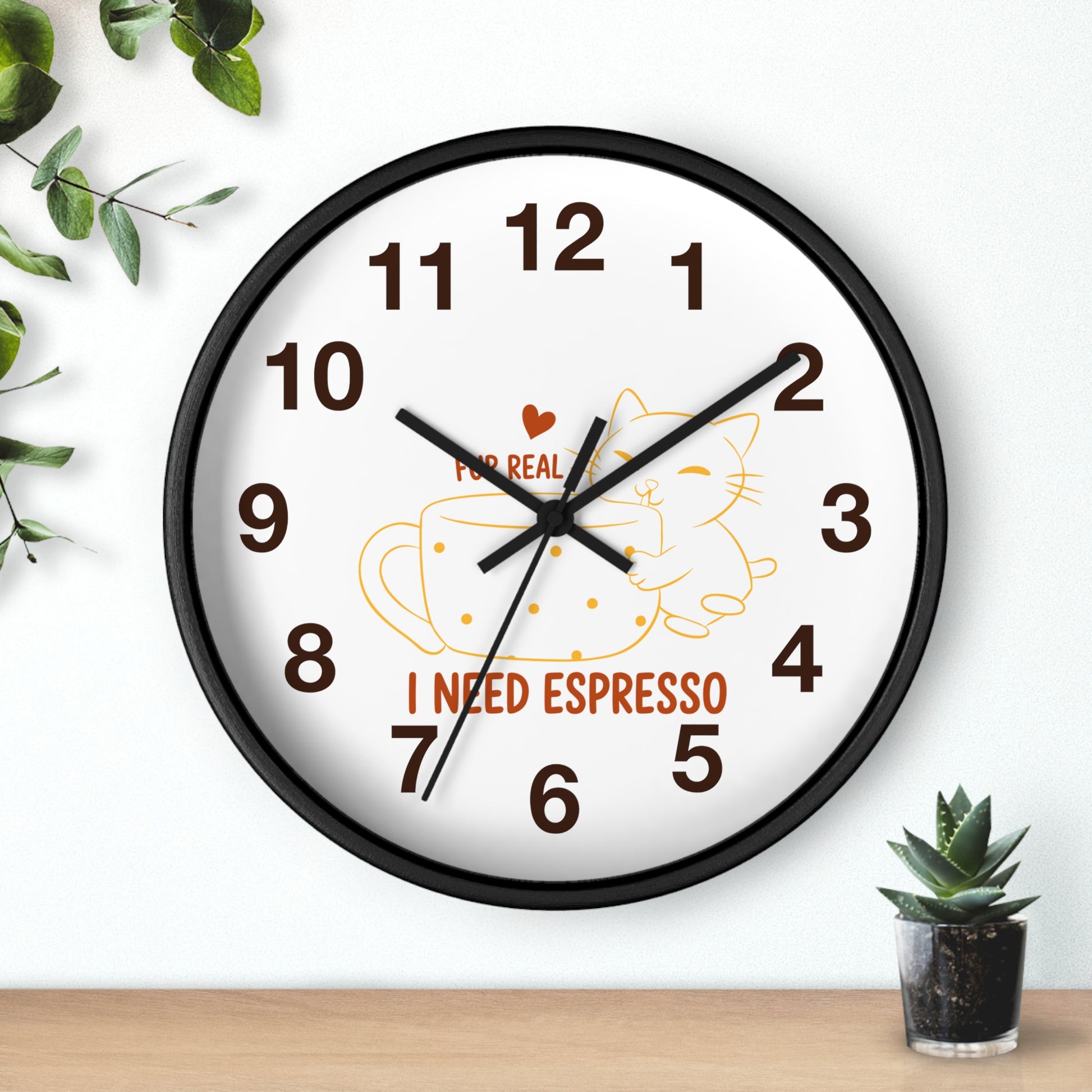 A round wall clock with a black frame and white face, featuring brown numbers, black hands, a yellow coffee cup and cat illustration, and the text 'I NEED ESPRESSO' with a small red heart. The clock is mounted on a white wall above a light wood surface, with green leaves and a small succulent nearby.