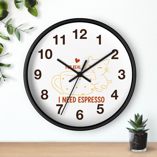 A round wall clock with a black frame and white face, featuring brown numbers, black hands, a yellow coffee cup and cat illustration, and the text 'I NEED ESPRESSO' with a small red heart. The clock is mounted on a white wall above a light wood surface, with green leaves and a small succulent nearby.