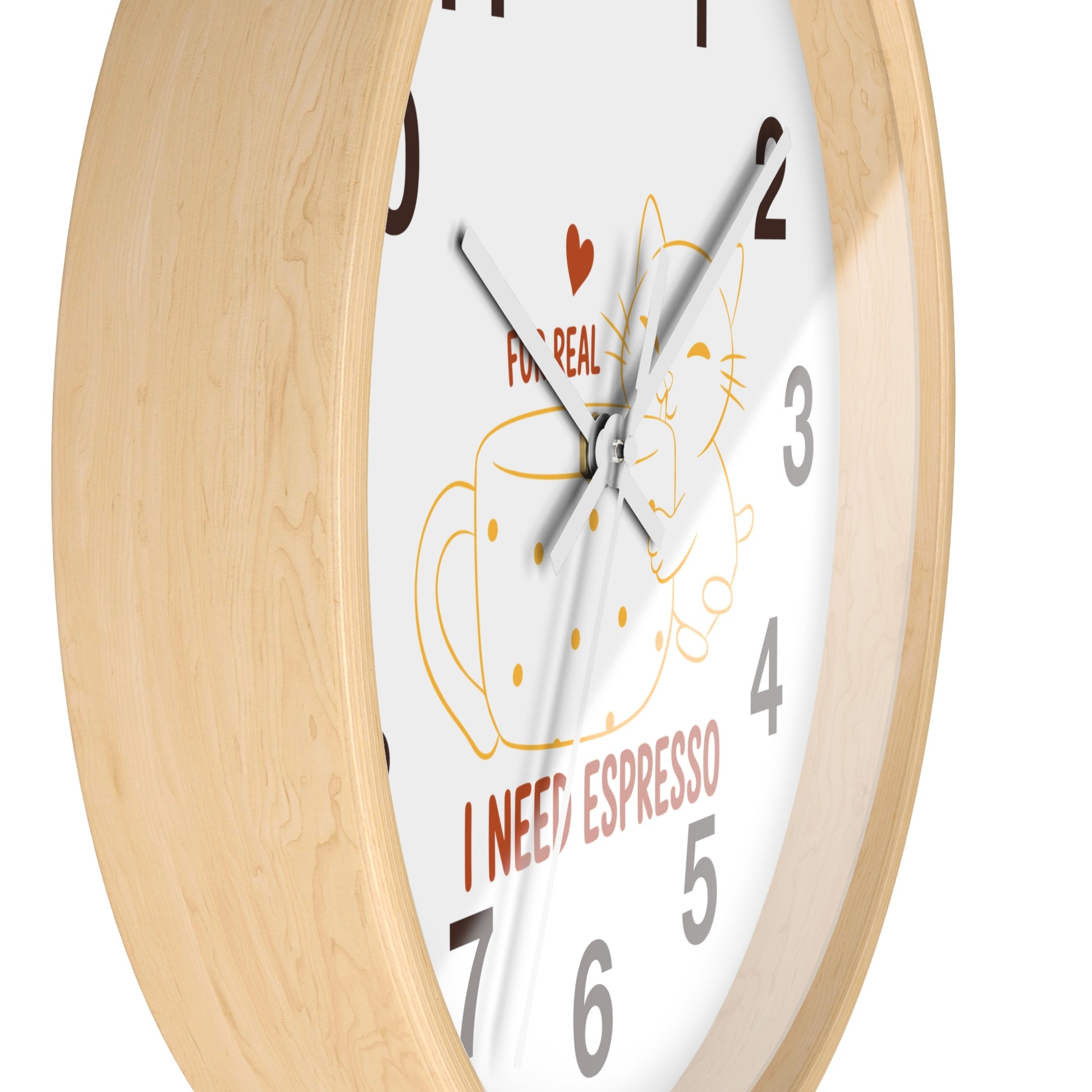 A close-up of a round wall clock with a light natural wood frame and a white face. The clock face features a whimsical illustration of a cat inside a dotted espresso cup, with the text 'FOR REAL I NEED ESPRESSO' and a small red heart.