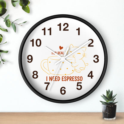 A round wall clock with a black frame and a white face, featuring a cartoon cat curled up inside an espresso cup with the text 'I NEED ESPRESSO' and a small red heart. The clock is shown hanging on a white wall above a wooden surface with small green plants.