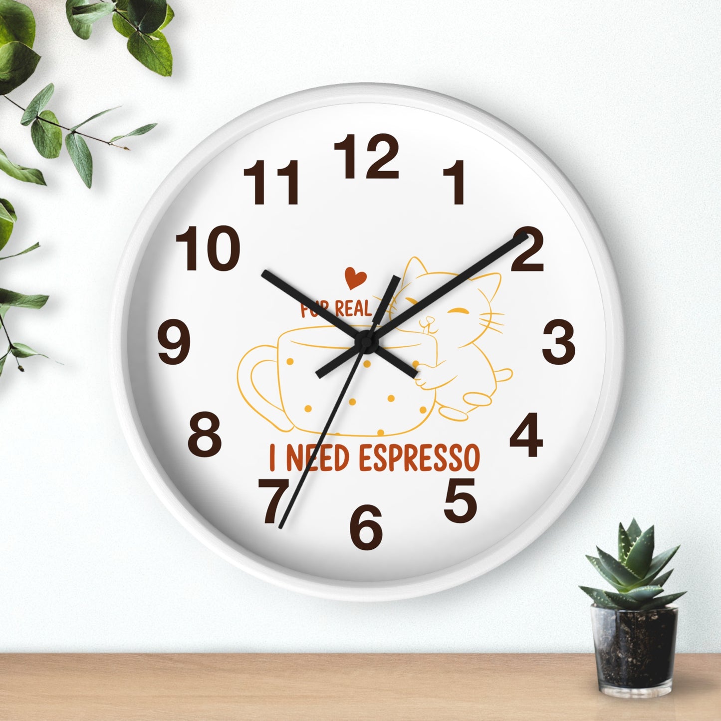 A round white wall clock with black hands and brown numerals, featuring a cartoon cat inside an espresso cup and the text 'I NEED ESPRESSO' on the face. The clock is shown hanging on a white wall above a wooden surface, with green leaves and a small potted succulent as decorative props.
