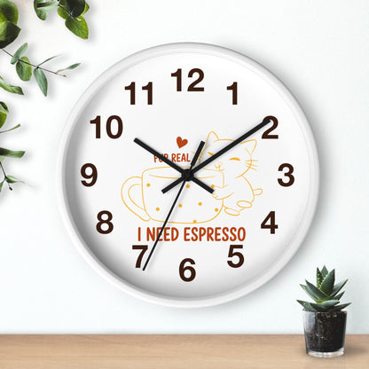 A round white wall clock with black hands and brown numerals, featuring a cartoon cat inside an espresso cup and the text 'I NEED ESPRESSO' on the face. The clock is shown hanging on a white wall above a wooden surface, with green leaves and a small potted succulent as decorative props.