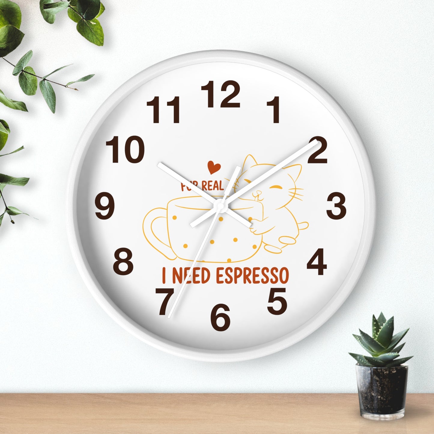 A round white wall clock with dark brown numbers and hands, featuring a cartoon cat inside an espresso cup and the words 'I NEED ESPRESSO' on the face. The clock is shown hanging on a white wall above a light wood surface, with decorative green leaves and a small potted succulent nearby.