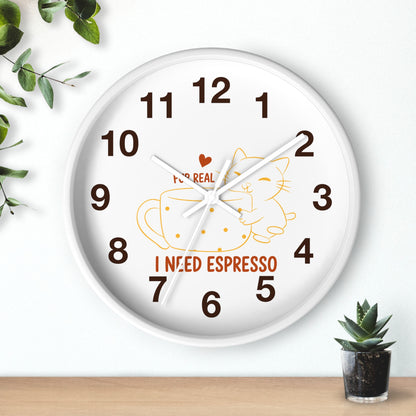 A round white wall clock with dark brown numbers and hands, featuring a cartoon cat inside an espresso cup and the words 'I NEED ESPRESSO' on the face. The clock is shown hanging on a white wall above a light wood surface, with decorative green leaves and a small potted succulent nearby.