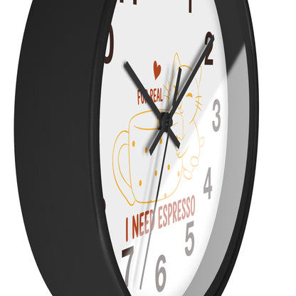 A close-up of a round wall clock with a black frame and white face. The clock face features a coffee cup illustration, a small red heart, the text 'FOR REAL', and 'I NEED ESPRESSO' in red near the bottom.