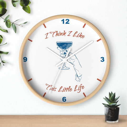 A round wall clock with a light wood frame and a white face, featuring the text 'I Think I Like This Little Life' in red script, blue numerals, and a blue illustration of a hand holding a cocktail glass. The clock is shown hanging on a white wall above a wooden surface with small green plants in the corners.