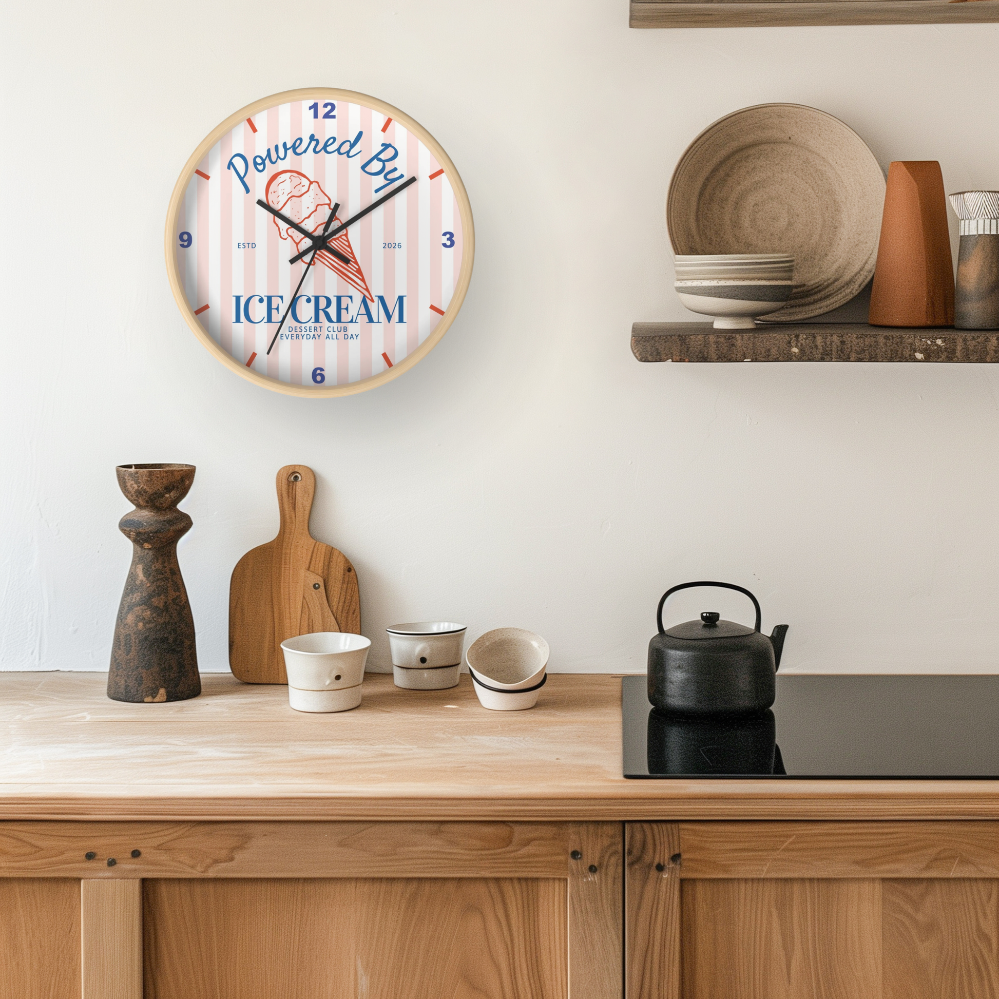 A round wall clock with a light wood frame hangs on a white wall above a wooden kitchen counter. The clock face has pink and white vertical stripes, blue text reading 'Powered By ICE CREAM', and a small ice cream cone illustration.