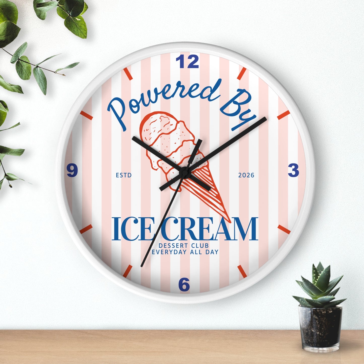 A round wall clock with a white frame and a light pink and white striped face. The clock face features a red and orange ice cream cone illustration, blue text reading 'Powered By ICE CREAM', and blue numerals for the hours.