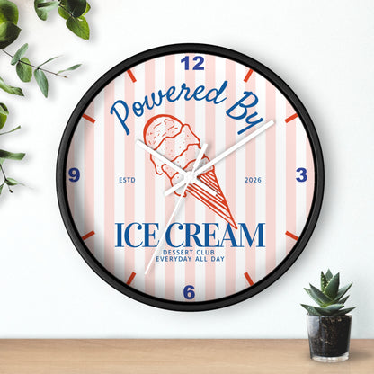 A round wall clock with a black frame and a white face featuring pink vertical stripes, blue text reading 'Powered By ICE cream CLUB,' and an ice cream cone illustration. The clock is shown hanging on a white wall above a wooden surface, with small green plants at the corners.