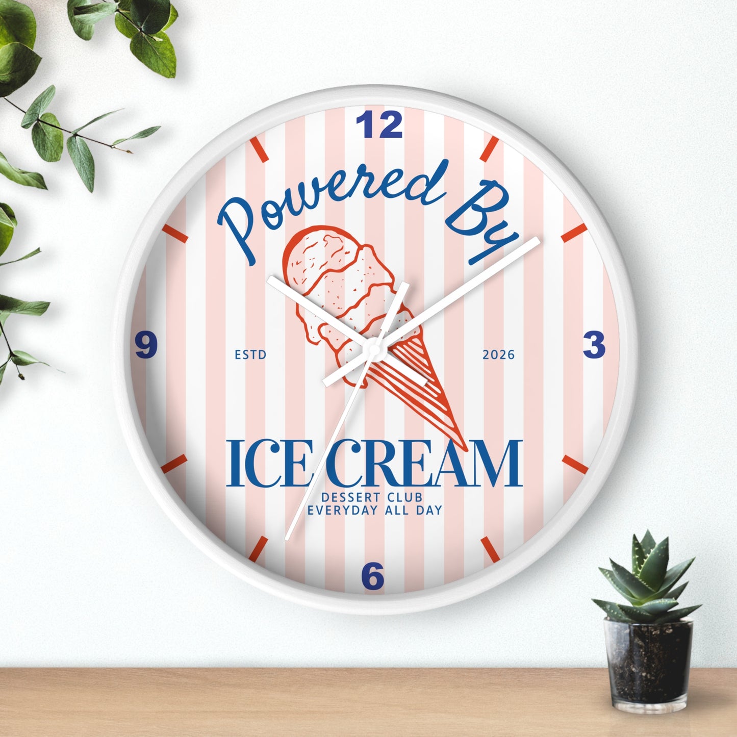 A round wall clock with a white frame and a pastel pink and white striped face. The clock features a red and orange ice cream cone illustration in the center, with the words 'Powered By' in blue script and 'ICE DREAM' in bold blue block letters.