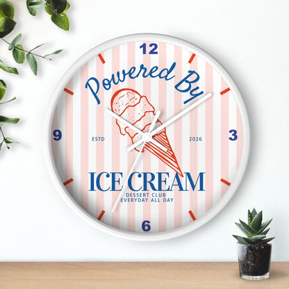 A round wall clock with a white frame and a pastel pink and white striped face. The clock features a red and orange ice cream cone illustration in the center, with the words 'Powered By' in blue script and 'ICE DREAM' in bold blue block letters.