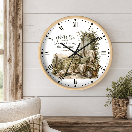 A round wall clock with a light wood frame hangs on a white shiplap wall above a wooden table. The clock face features a vintage illustration of a porch with a rocking chair, flowers, and a fence, with the word 'grace' and the phrase 'steps through the waiting' in script and serif fonts.