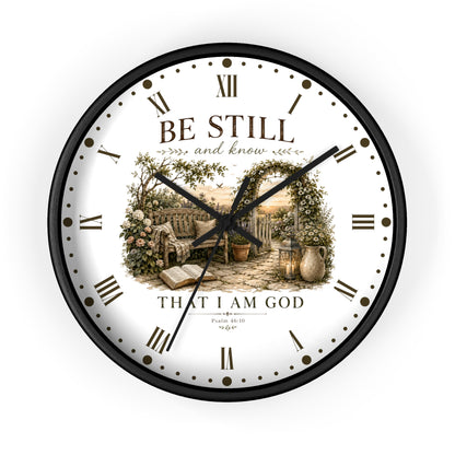 A round wall clock with a black frame and a white face featuring a vintage-style illustration of a garden scene with an open book and the text 'BE STILL and know THAT I AM GOD' with scripture reference Philippians 4:13.