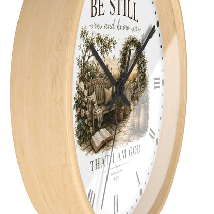 A round wall clock with a light natural wood frame and a white face featuring a vintage-style illustration of a garden bench, flowers, and an open book, with the words 'BE STILL and know' and 'THAT I AM GOD' on a white background.