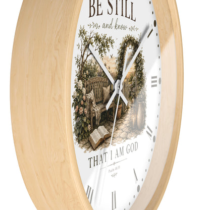 A round wall clock with a light natural wood frame and a white face featuring a vintage-style illustration of a garden bench under an arched trellis, with the text 'BE STILL and know' and 'THAT I AM GOD'.