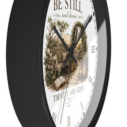 A round wall clock with a black frame and a white face featuring a vintage-style illustration of a garden bench, flowers, and an open book, with the text 'BE STILL and know' and 'THAT I AM GOD' on the dial.