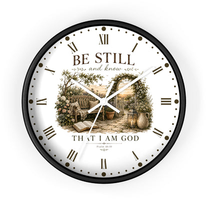 A round wall clock with a black frame and a white face featuring the text 'BE STILL and know THAT I AM GOD' and a vintage-style garden scene in the center, shown on a plain white background.