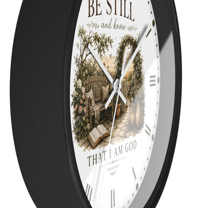 A round wall clock with a black frame and a white face featuring a vintage-style illustration of a garden bench beneath an arched floral trellis, with the text 'BE STILL and know' and 'THAT I AM GOD' on a white background.