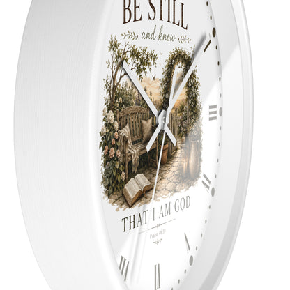 A white round wall clock with a vintage-style printed illustration of a garden bench under an arched trellis and the words 'BE STILL and know' and 'THAT I AM GOD' on the dial.