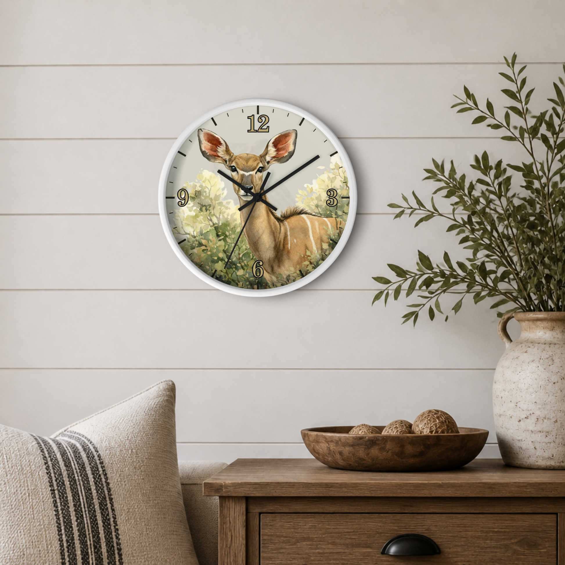 A round wall clock with a white frame and a deer illustration on the face, mounted on a light wood shiplap wall above a wooden side table with a bowl and a vase of greenery.