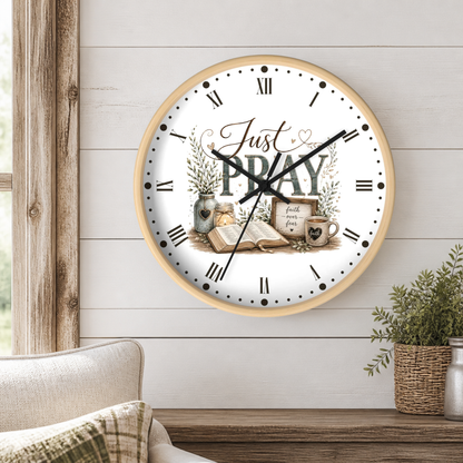 A round wall clock with a light wood frame hangs on a white shiplap wall above a wooden table. The clock face is white with black Roman numerals and hands, featuring the words 'Just PRAY' and a vintage-style illustration of an open book, a mug, and small plants.