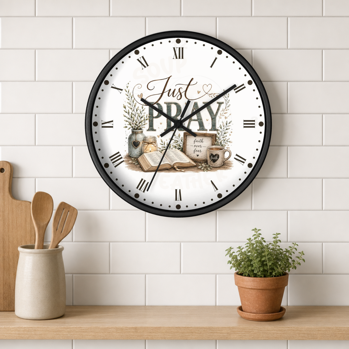 A round wall clock with a black frame and a white face featuring the words 'Just PRAY' and a rustic decorative illustration, mounted on a white subway tile wall above a wooden shelf with kitchen utensils and a small potted plant.