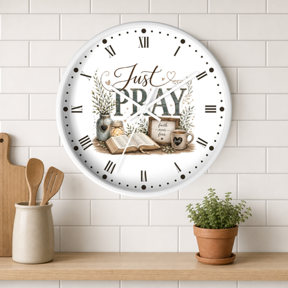 A round white wall clock with Roman numerals and a decorative religious illustration reading 'Just PRAY' in a kitchen setting, mounted on a white subway tile wall above a wooden shelf with utensils and a potted plant.