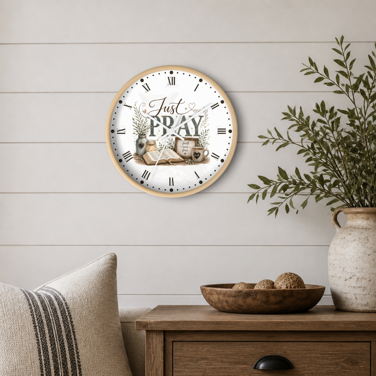 A round wall clock with a light wood frame and a white face featuring the words 'Just PRAY' in script and block lettering, styled on a white shiplap wall above a wooden side table with a vase and greenery.