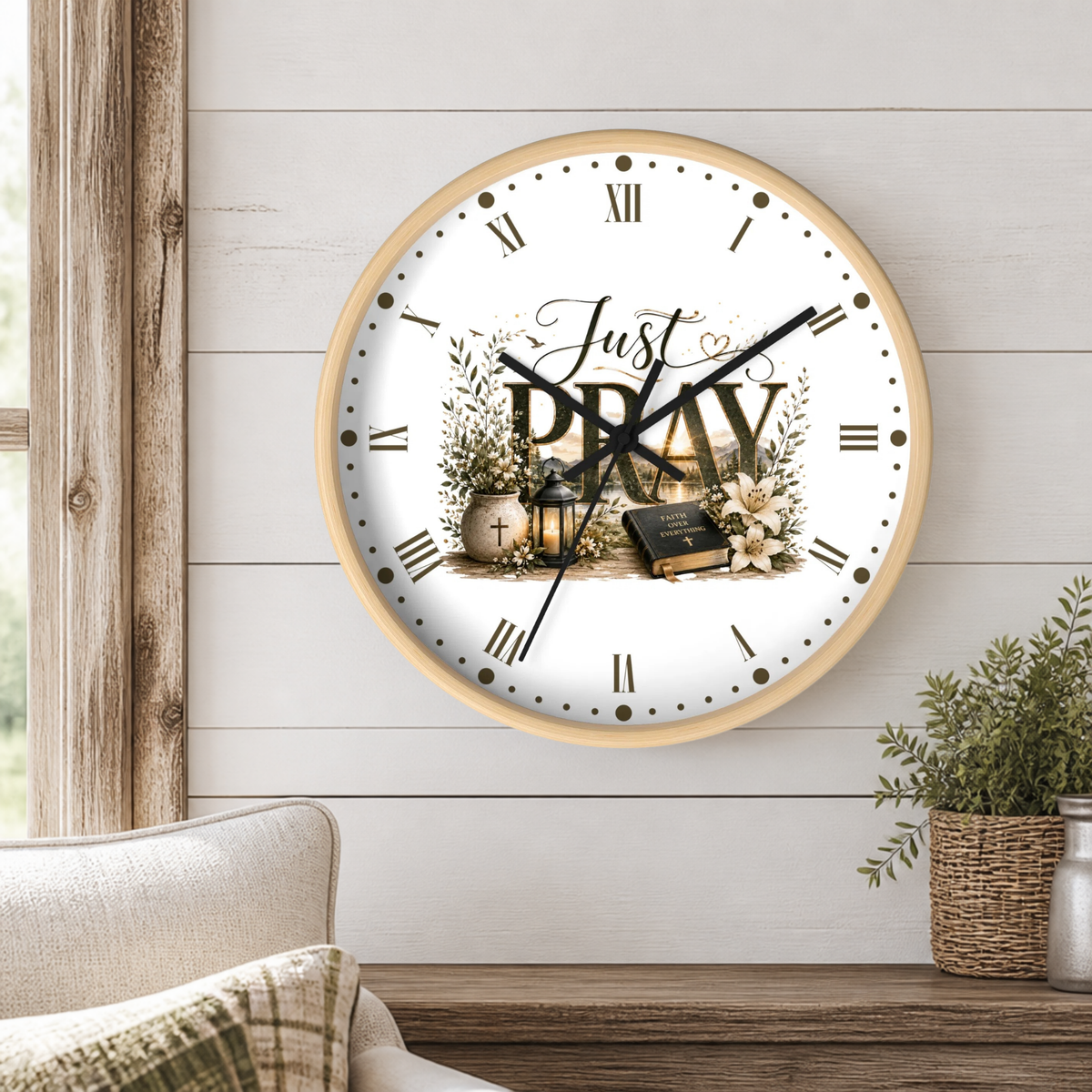 A round wall clock with a light wood frame hangs on a white shiplap wall above a wooden table. The clock face is white with black Roman numerals, black hands, and a central graphic featuring the words 'Just PRAY' surrounded by floral illustrations, a cross, and a Bible.