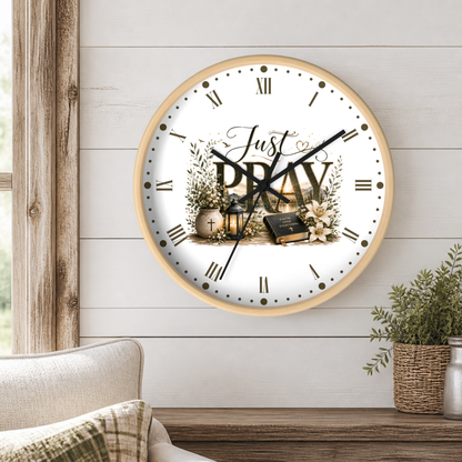 A round wall clock with a light wood frame hangs on a white shiplap wall above a wooden table. The clock face is white with black Roman numerals, black hands, and a central graphic featuring the words 'Just PRAY' surrounded by floral illustrations, a cross, and a Bible.