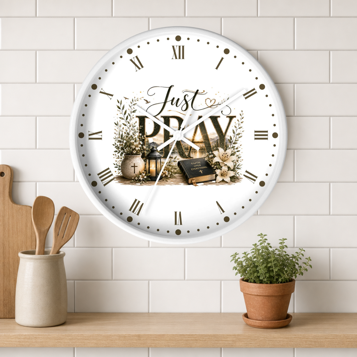 A round white wall clock with Roman numerals and the words 'Just PRAY' in the center, styled on a white subway tile wall above a wooden shelf with kitchen utensils and a potted plant.