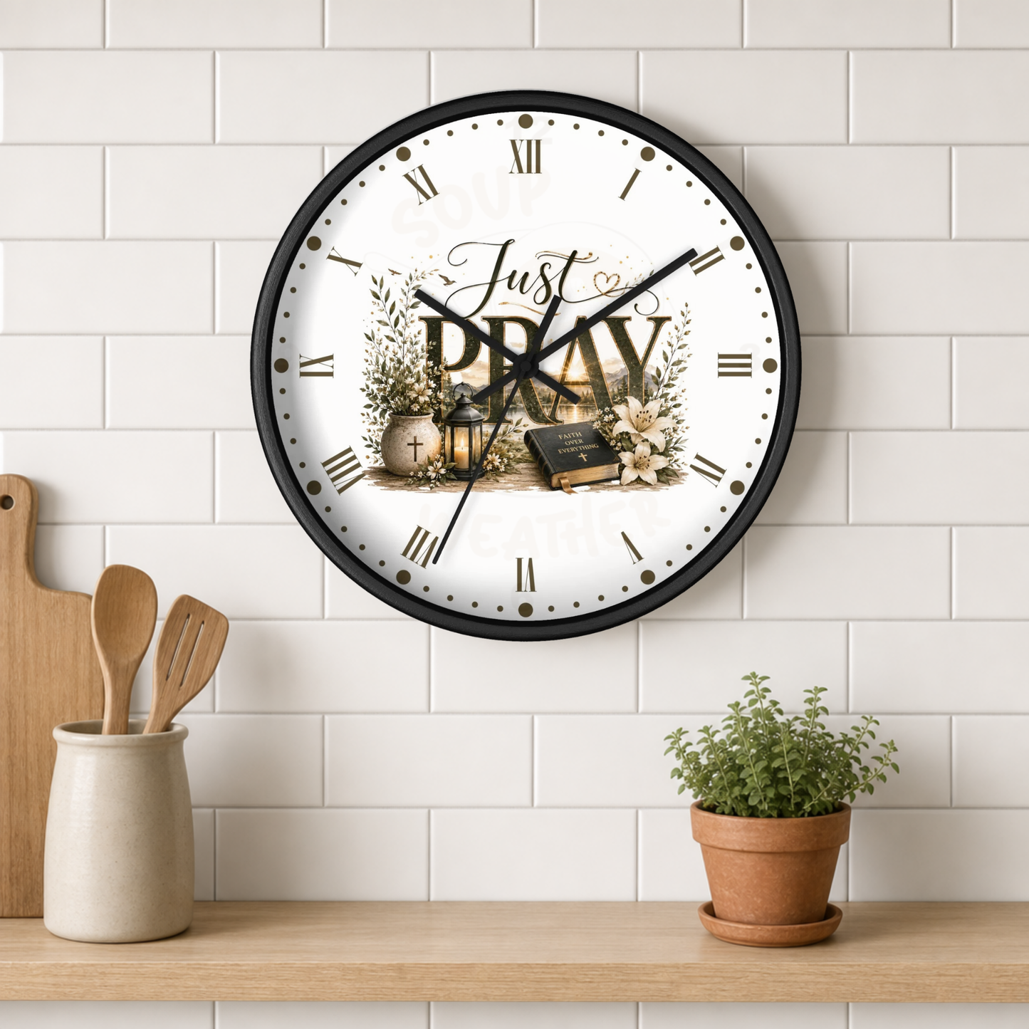 A round wall clock with a black frame and a white face featuring the words 'Just PRAY' with religious illustrations, mounted on a white subway tile wall above a wooden shelf with kitchen utensils and a small potted plant.