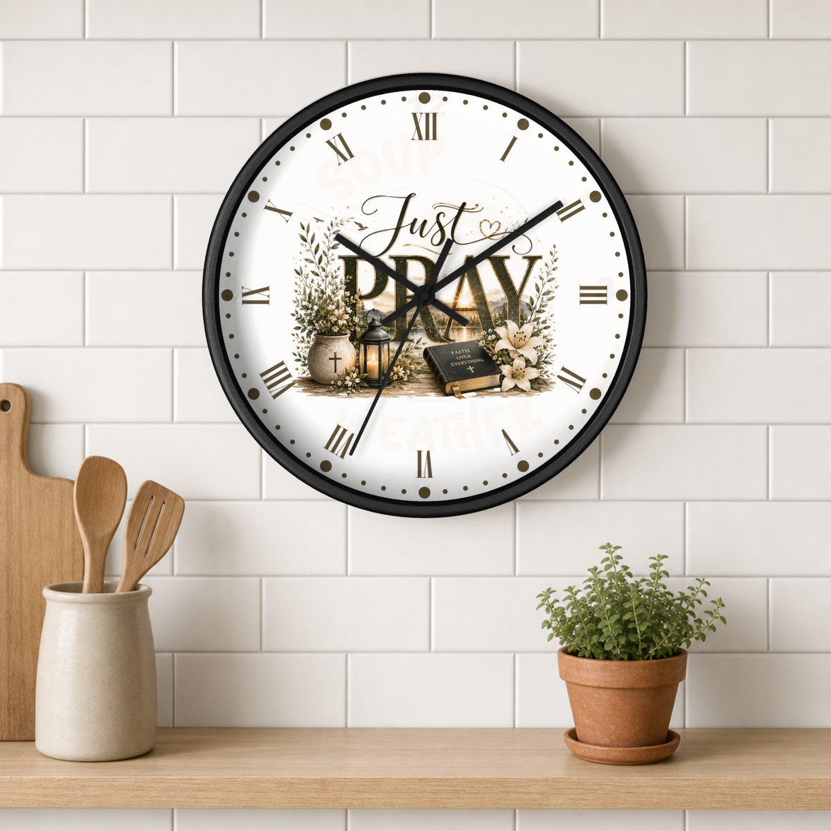 A round wall clock with a black frame and a white face featuring the words 'Just PRAY' with religious illustrations, mounted on a white subway tile wall above a wooden shelf with kitchen utensils and a small potted plant.