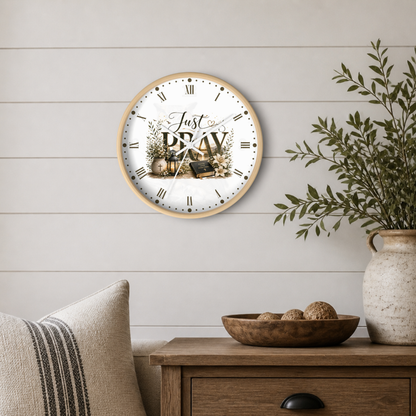 A round wall clock with a light wood frame and a white face featuring the words 'Just PRAY' with a small house, lantern, and floral design. The clock is mounted on a white shiplap wall above a wooden side table with a bowl, decorative pillow, and a potted green plant in a ceramic vase.