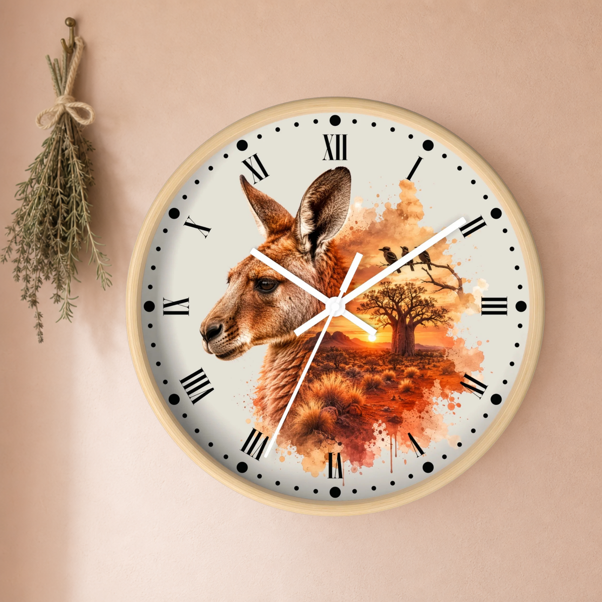 A round wall clock with a light wood frame and a printed design featuring a kangaroo head over an autumn landscape with a tree and sunset. The clock is mounted on a beige wall, with a dried herb sprig hanging on the left.