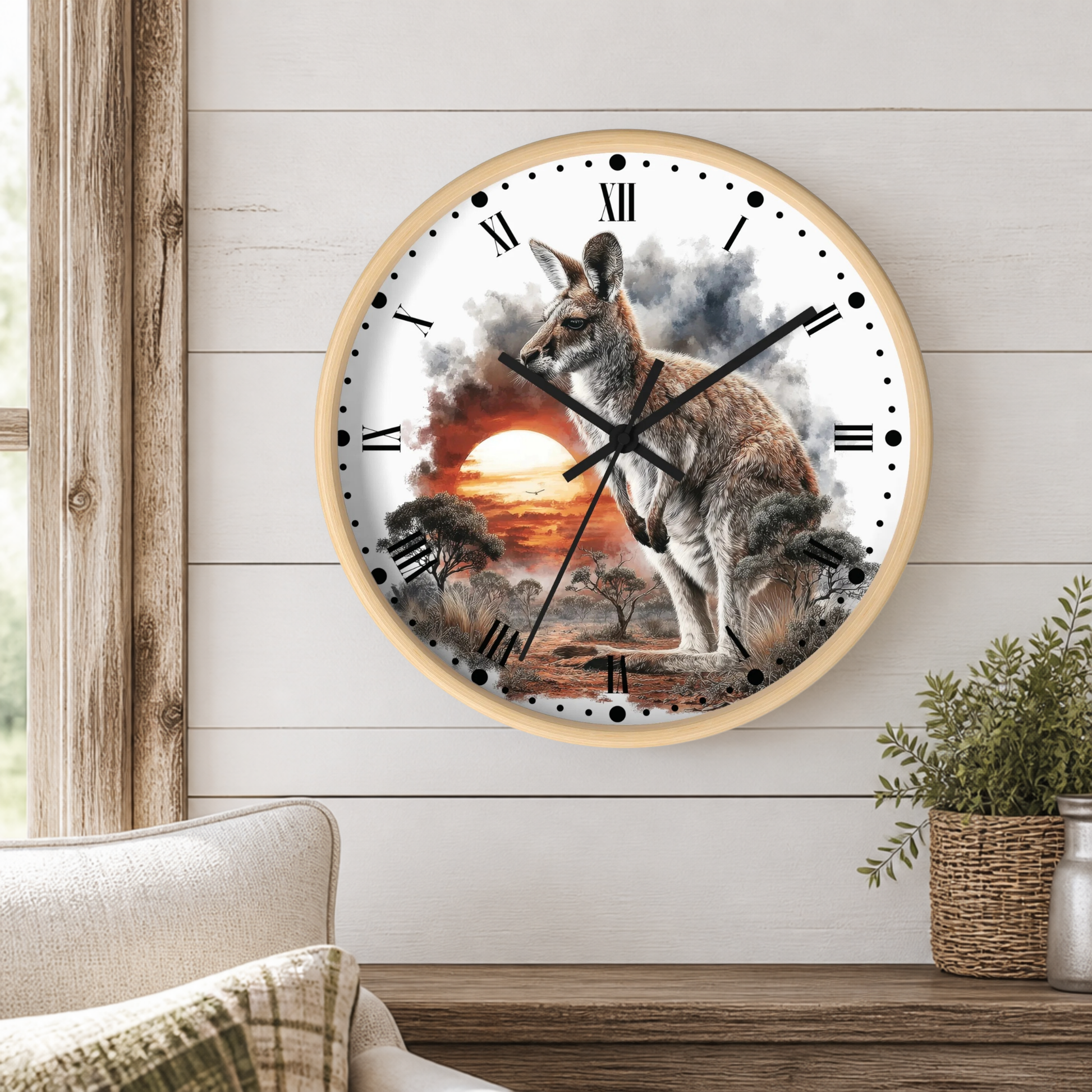 A round wall clock with a light wood frame hangs on a white shiplap wall above a wooden table. The clock face features a detailed illustration of a kangaroo standing in a sunset landscape with smoke and clouds.