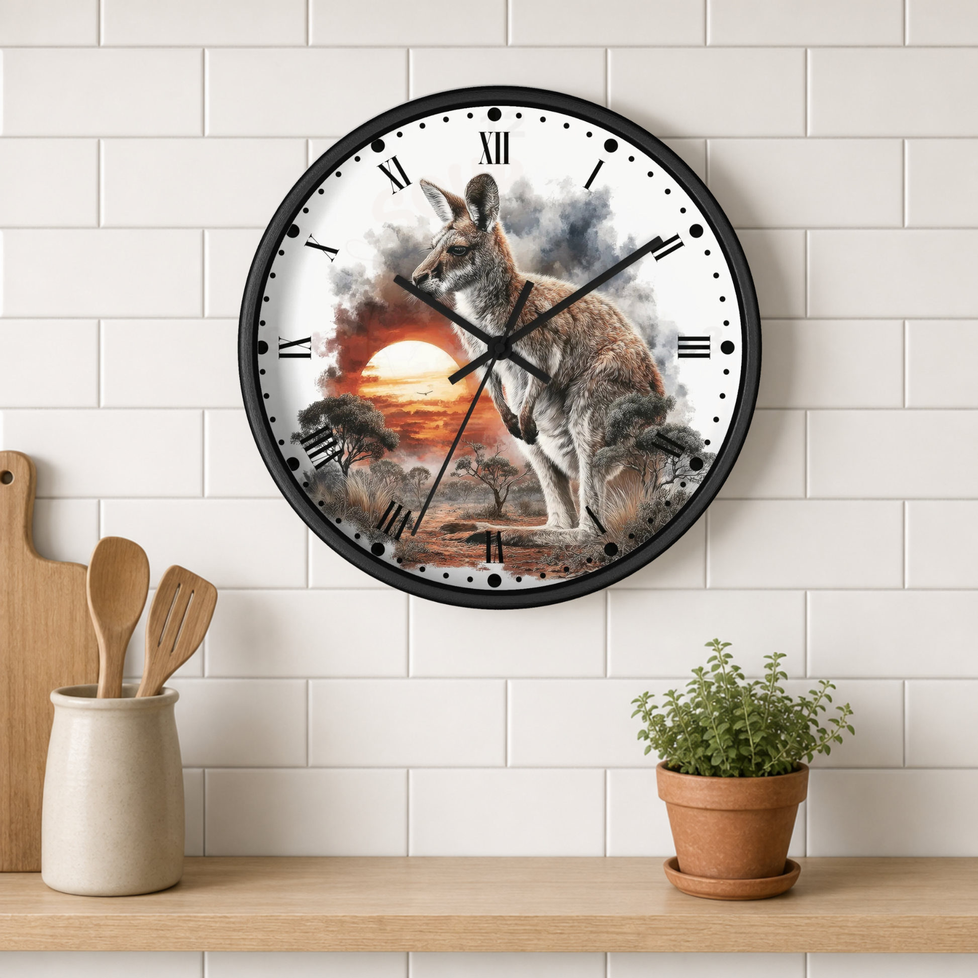 A round wall clock with a black frame hangs on a white subway tile wall above a wooden shelf. The clock face features a detailed illustration of a kangaroo standing in a sunset landscape.