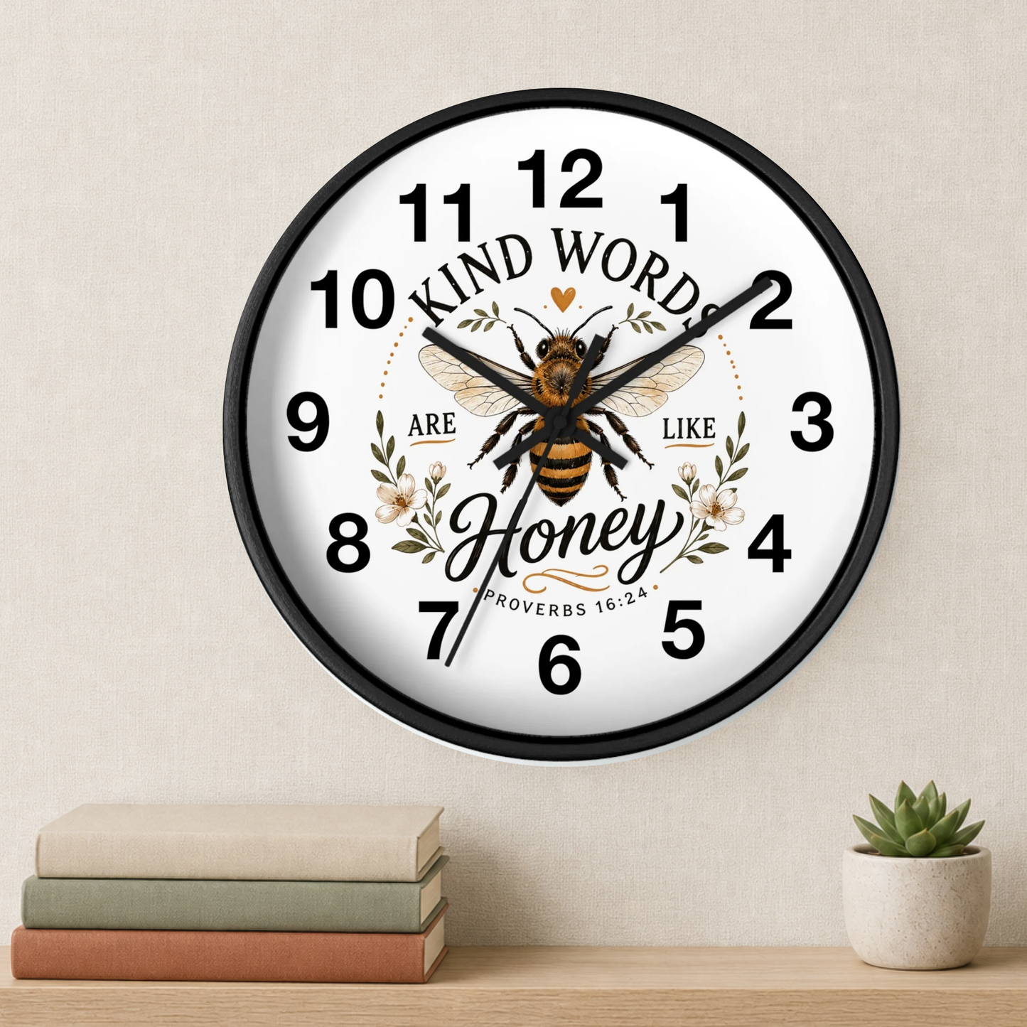 A round wall clock with a black frame and a white face, featuring a bee illustration and the words 'Kind Words Are Like Honey' with floral accents. The clock is mounted on a beige wall above a wooden shelf with stacked books and a small potted succulent.