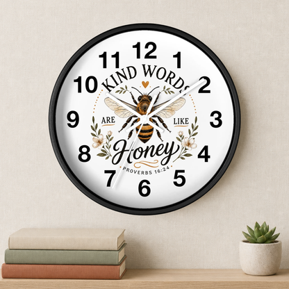 A round wall clock with a black frame and a white face featuring a bee illustration and the words 'Kind Words Are Like Honey' with a Bible verse. The clock is mounted on a beige wall above a shelf with stacked books and a small potted succulent.