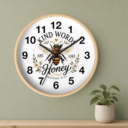 A round wall clock with a light wood frame and a white face, featuring a honeybee illustration and the words 'KIND WORD ARE LIKE Honey' with floral accents. The clock is mounted on a sage green wall above a wooden shelf with a small potted plant.