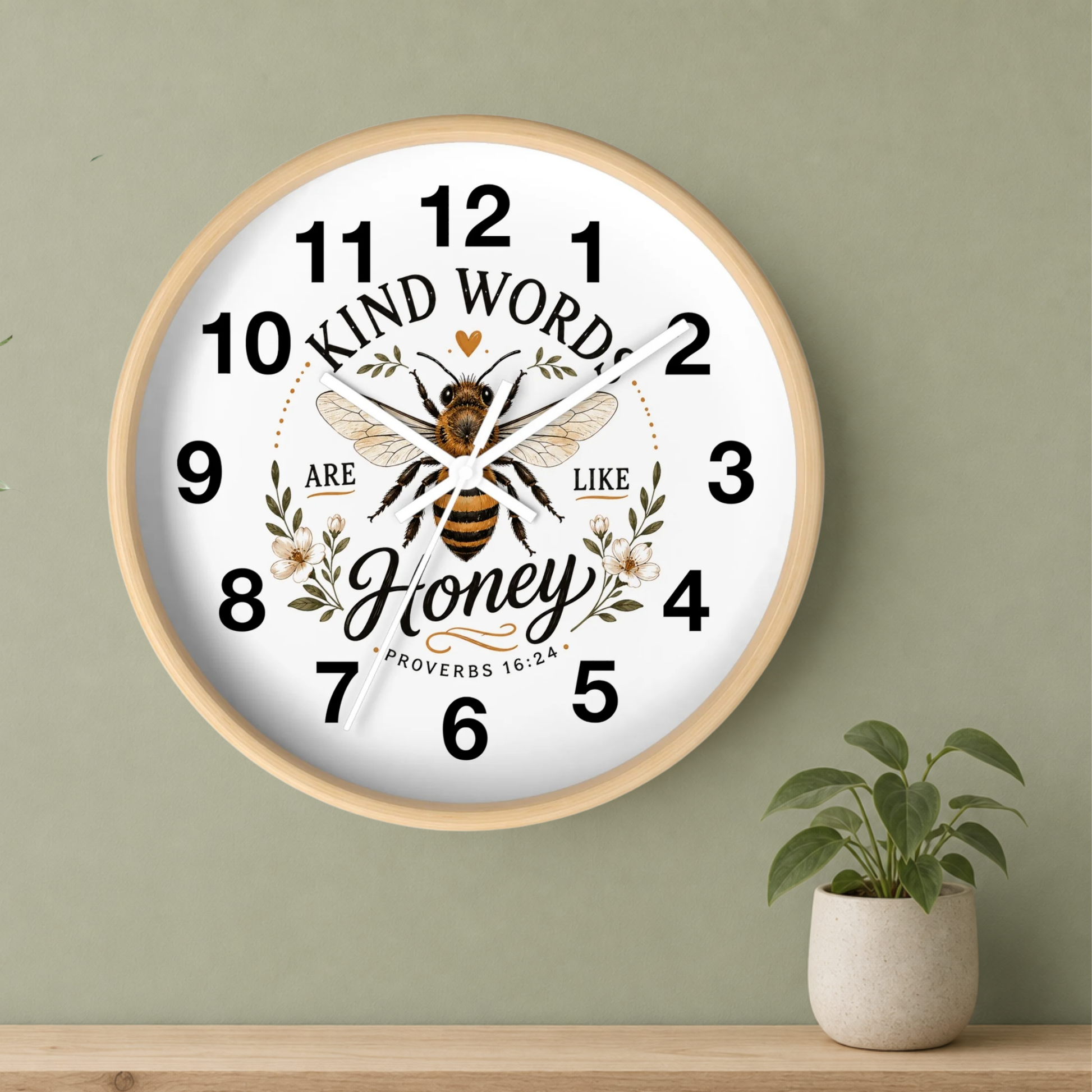 A round wall clock with a light wood frame and a white face, featuring a bee illustration and the text 'KIND WORDS ARE LIKE Honey PROVERBS 16:24.' The clock is mounted on a sage green wall above a wooden shelf with a small potted plant.