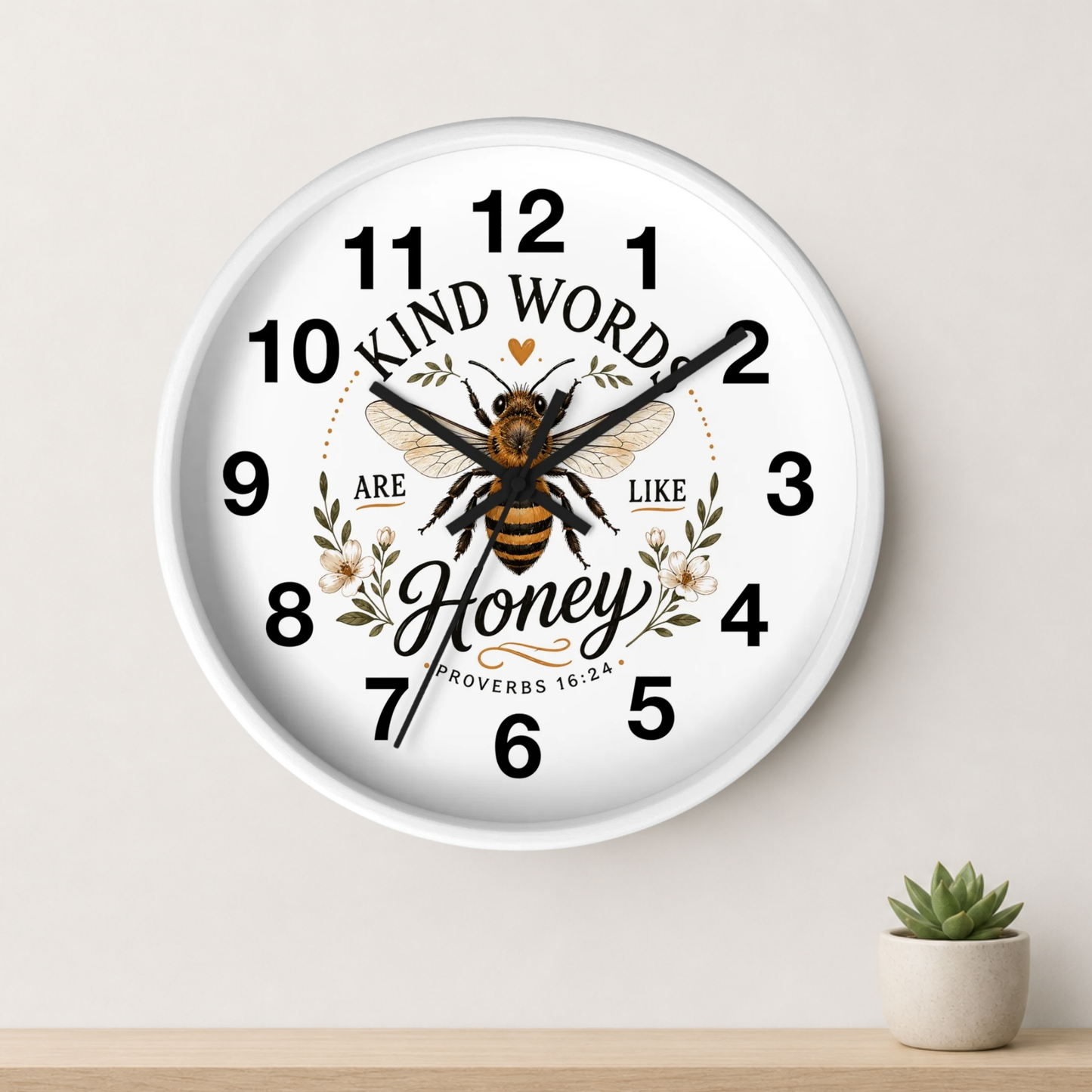 A round white wall clock with a printed bee, floral details, and the words 'KIND WORDS ARE LIKE Honey' on the face, shown hanging on a light wall above a small potted succulent.